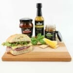 Our grazing board gift set includes a wooden board, chutney, chili sauce, and pesto, pictured along with a sandwich.