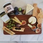 This charcuterie board gift set includes a charcuterie board, fig jam, and olive tapenade, shown with cheese, fresh figs, crackers, and olives.