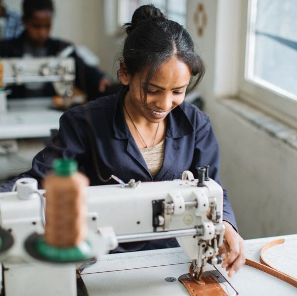 The leather backpack for travel is made by ABLE, a company that provides living wages and healthcare to workers like this woman shown sitting at a sewing machine.