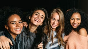 A group of women smile with their arms aorund each other. This list of women's day gift ideas for employees includes gifts from women-owned and women-led organizations.