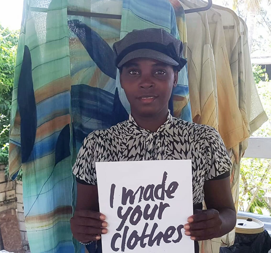 An artist holds up a sign reading "I made your clothes" in front of finished fashion pieces