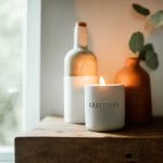 Lit gratitude candle on a table in front of a bottle and vase